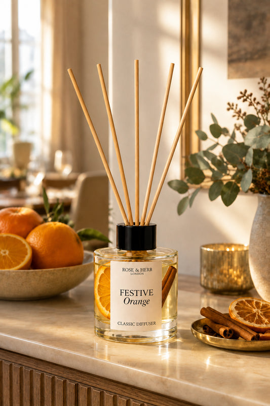 A ROSE & HERB London 'Festive Orange' classic reed diffuser bottle with a black cap, filled with amber liquid and orange slices, surrounded by natural reed sticks. The diffuser sits on a marble countertop in a warm, sunlit room with oranges, cinnamon sticks, and greenery in the background.