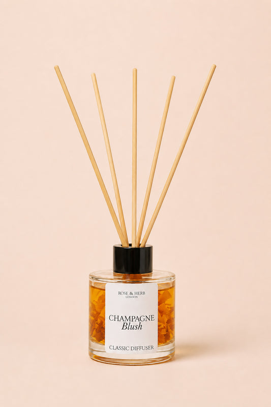 A reed diffuser bottle with a black cap and natural wood reeds, filled with amber liquid, labeled 'Champagne Blush' by Rose & Herb, set against a solid pale peach background.