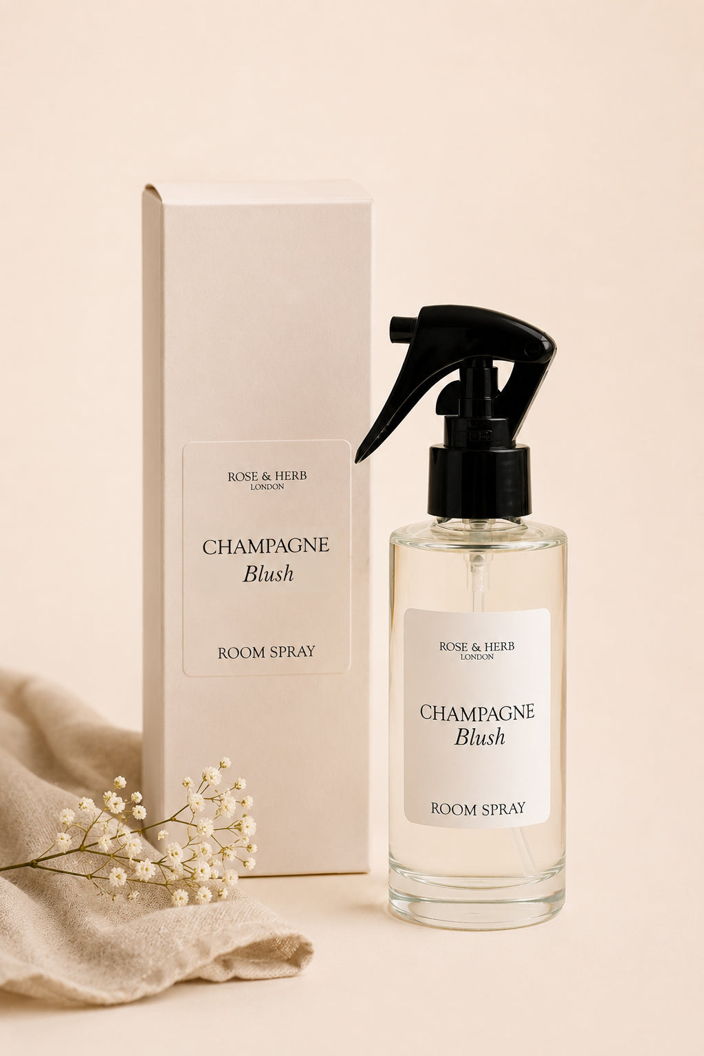 A bottle of Rose & Herb London Champagne Blush room spray next to its cream-colored packaging box, styled with a beige fabric and small white baby's breath flowers on a warm beige background.