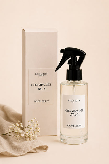 A bottle of Rose & Herb London Champagne Blush room spray next to its cream-colored packaging box, styled with a beige fabric and small white baby's breath flowers on a warm beige background.