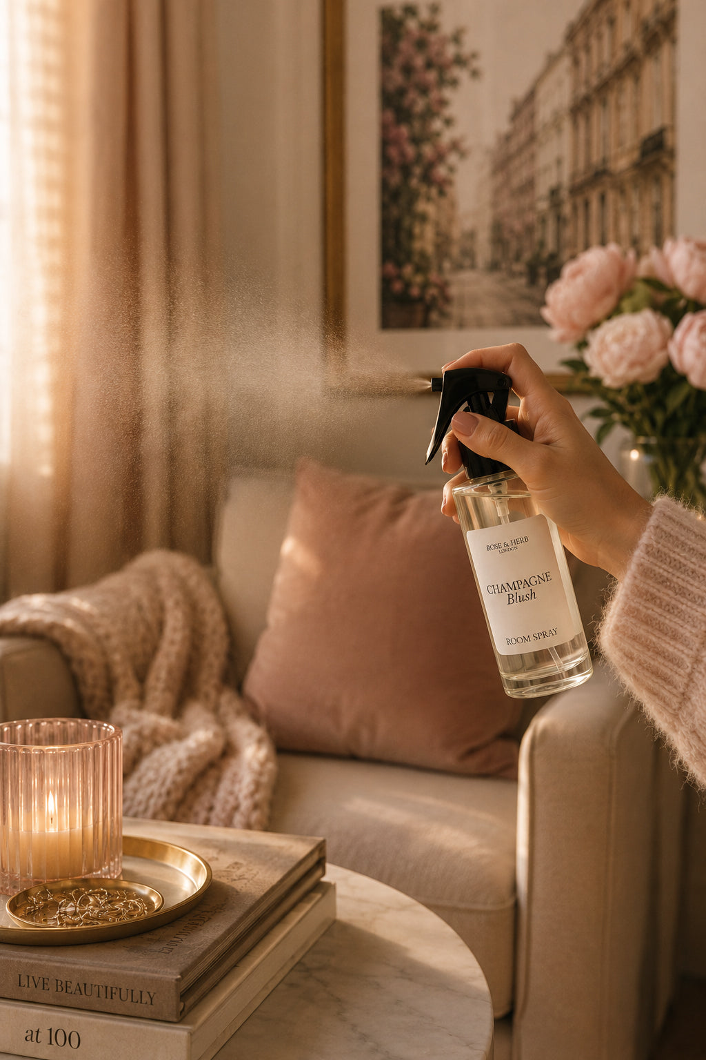 A hand holds a clear spray bottle labeled 'Champagne Blush' in a warm, cozy living room setting. The scene includes a beige sofa with a pink pillow, a lit candle on stacked books, and pink flowers in the background.