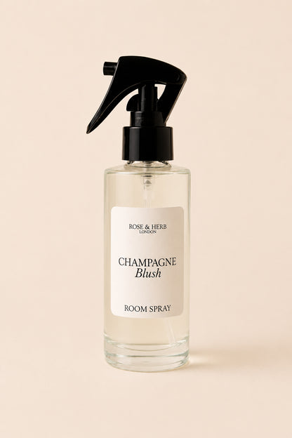 A clear glass room spray bottle with a black trigger spray top and a cream label reading 'Champagne Blush Room Spray' by Rose & Herb London, set against a solid pale peach background.