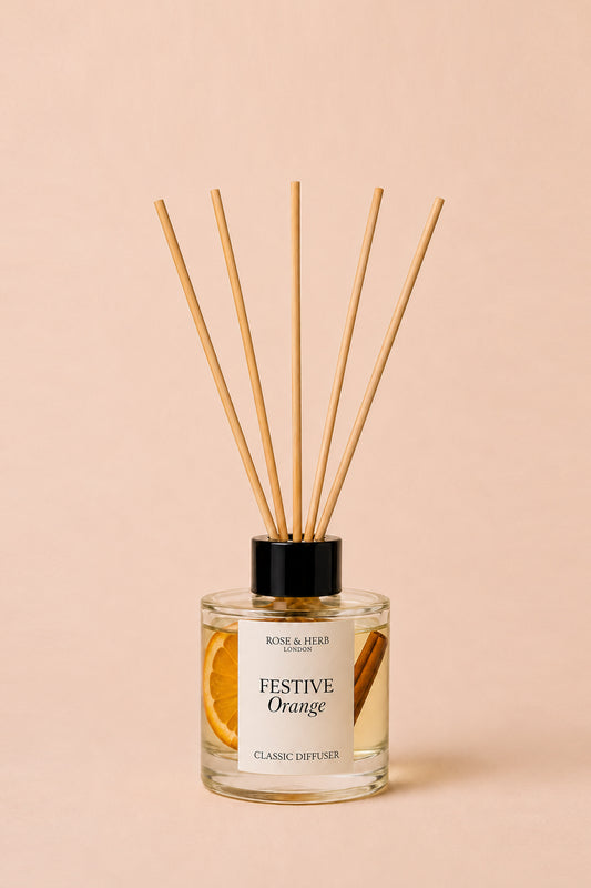 A reed diffuser bottle with clear glass, amber liquid, a black cap, and six natural reed sticks, labeled 'Festive Orange' by Rose & Herb Scents, set against a solid pale peach background.