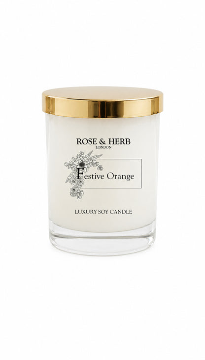 A Rose & Herb London 'Festive Orange' luxury soy candle in a clear glass jar with a shiny gold lid, set against a plain white background.