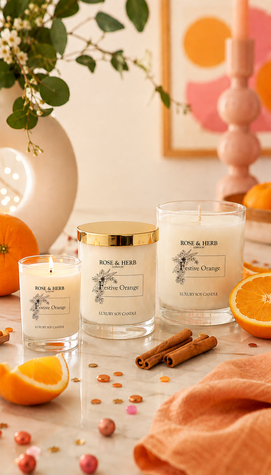 Three ROSE & HERB scented candles in glass jars with white wax are styled on a marble surface with orange slices, cinnamon sticks, and a peach fabric, against a warm, softly blurred background.