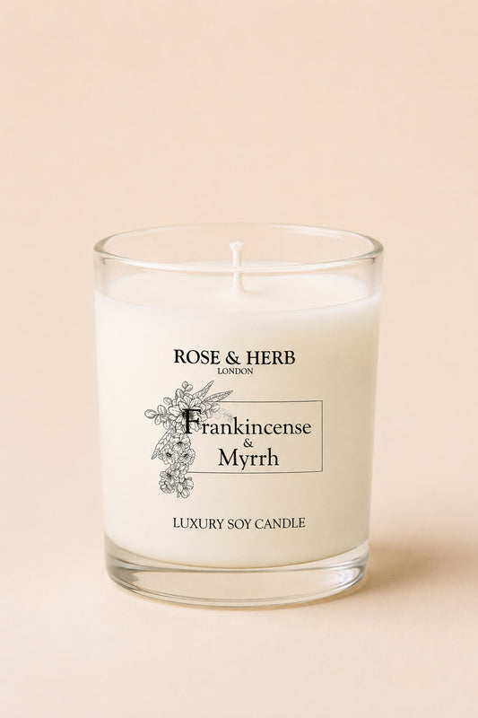 A white soy candle in a clear glass jar with a printed label reading 'ROSE & HERB LONDON' and 'Frankincense & Myrrh', set against a solid pale peach background.
