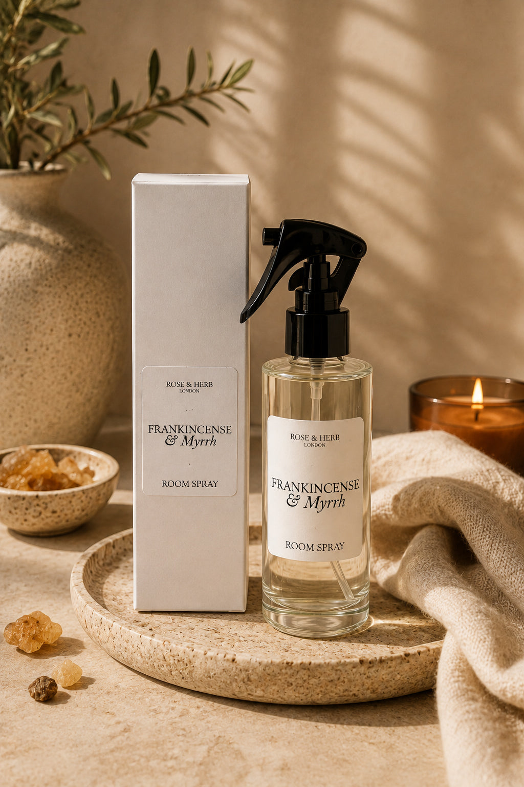 A room spray bottle and its matching box displayed on a round tray, styled with a lit candle, beige fabric, and dried botanicals in a warm, sunlit setting.