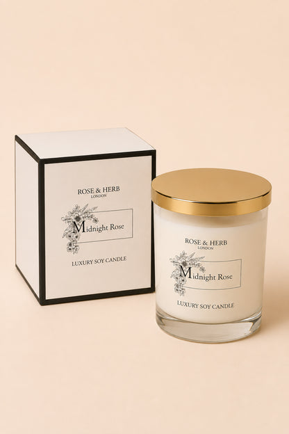 A luxury soy candle in a clear glass jar with a gold lid, shown next to its white and black box packaging. The labels read 'ROSE & HERB LONDON' and 'Midnight Rose'.