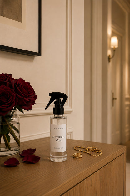 A clear glass spray bottle labeled 'Midnight Rose' by ROSE & HERB, placed on a wooden console table next to a vase of deep red roses and gold rings in a warm, elegant interior setting.