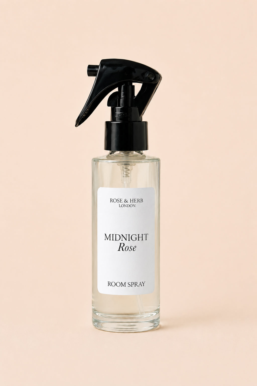 A clear glass room spray bottle with a black trigger sprayer and a white label reading 'MIDNIGHT Rose' and 'ROSE & HERB LONDON', centered on a soft peach background.