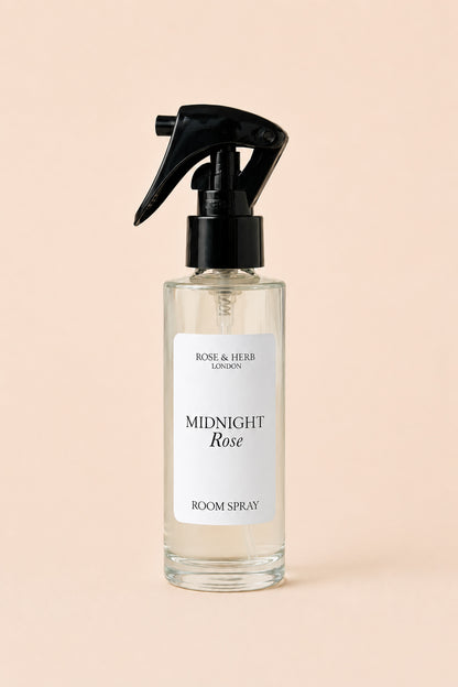 A clear glass room spray bottle with a black trigger sprayer and a white label reading 'MIDNIGHT Rose' and 'ROSE & HERB LONDON', centered on a soft peach background.