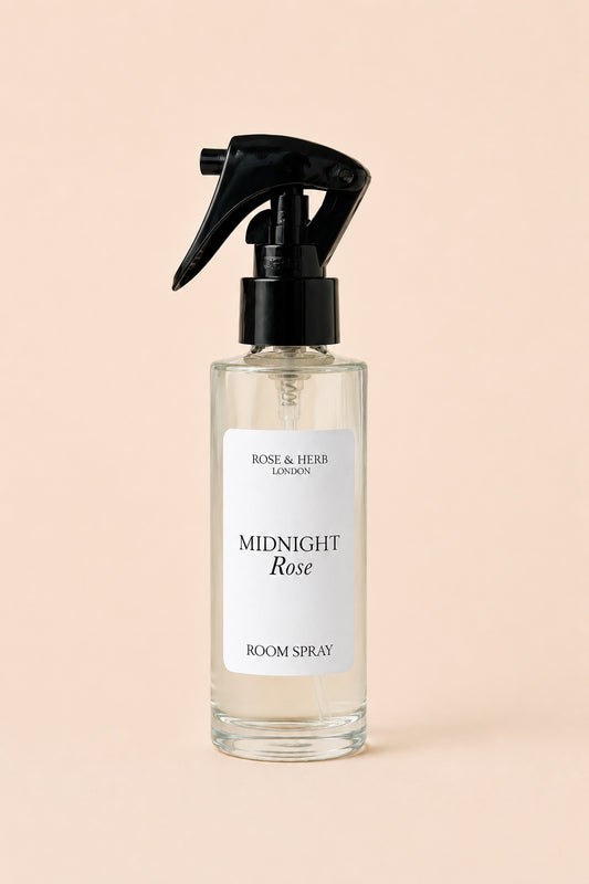 A clear glass room spray bottle with a black trigger sprayer and a white label reading 'MIDNIGHT Rose' and 'ROSE & HERB LONDON', centered on a soft peach background.