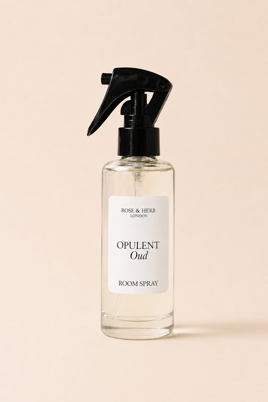 A clear glass room spray bottle with a black trigger sprayer and a white label reading 'OPULENT Oud' and 'ROSE & HERB LONDON', centered on a warm beige background.