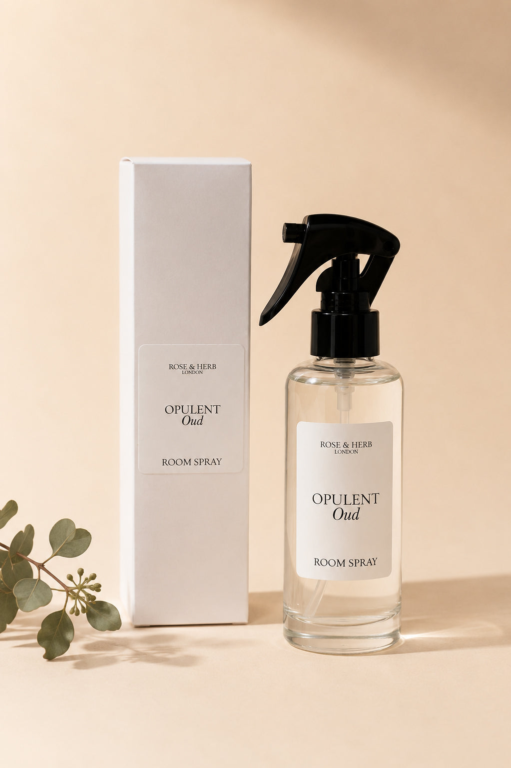 A clear glass room spray bottle with a black trigger sprayer and a white label reading 'OPULENT Oud ROOM SPRAY' by Rose & Tier, shown beside its tall white box on a warm beige background with a small green branch at the lower left.