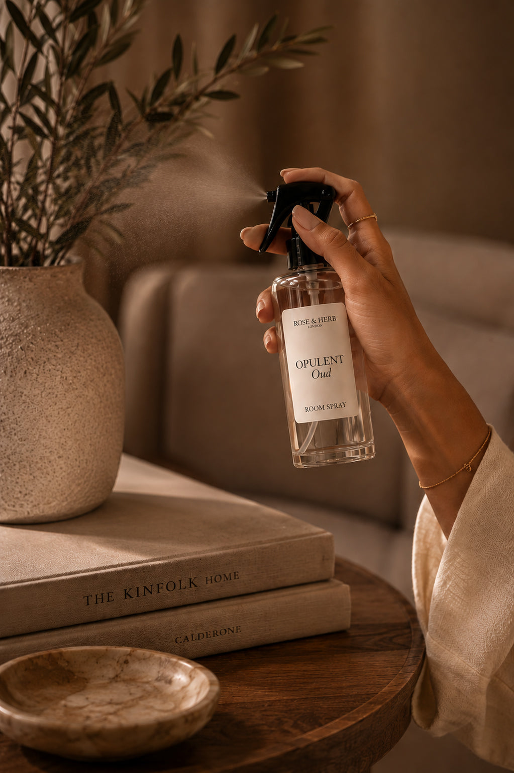 A hand holds a clear glass spray bottle labeled 'OPULENT Oud' by Rose Albers, misting a room with warm ambient light. The bottle is shown in a cozy interior setting with a wooden table, books, and a vase in the background.