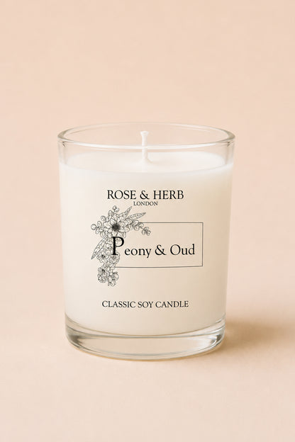 A classic soy candle in a clear glass jar with white wax, featuring a black printed label that reads 'ROSE & HERB LONDON' and 'Peony & Oud'. The candle is set against a solid, warm beige background.