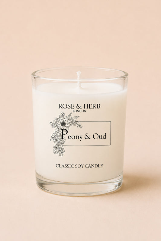 A classic soy candle in a clear glass jar with white wax, featuring a black printed label that reads 'ROSE & HERB LONDON' and 'Peony & Oud'. The candle is set against a solid, warm beige background.