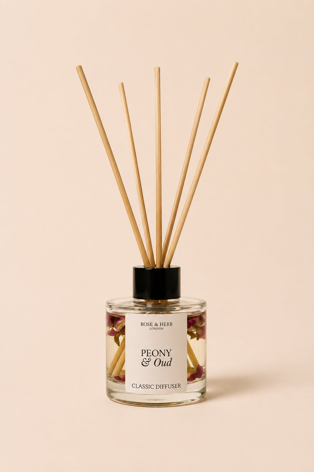 A reed diffuser bottle with a black cap and natural wooden reeds, labeled 'PEONY & Oud' by Rose & Herb, set against a solid pale peach background.
