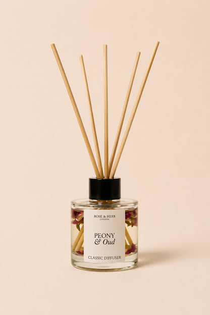 A reed diffuser bottle with a black cap and natural wooden reeds, labeled 'PEONY & Oud' by Rose & Herb, set against a solid pale peach background.