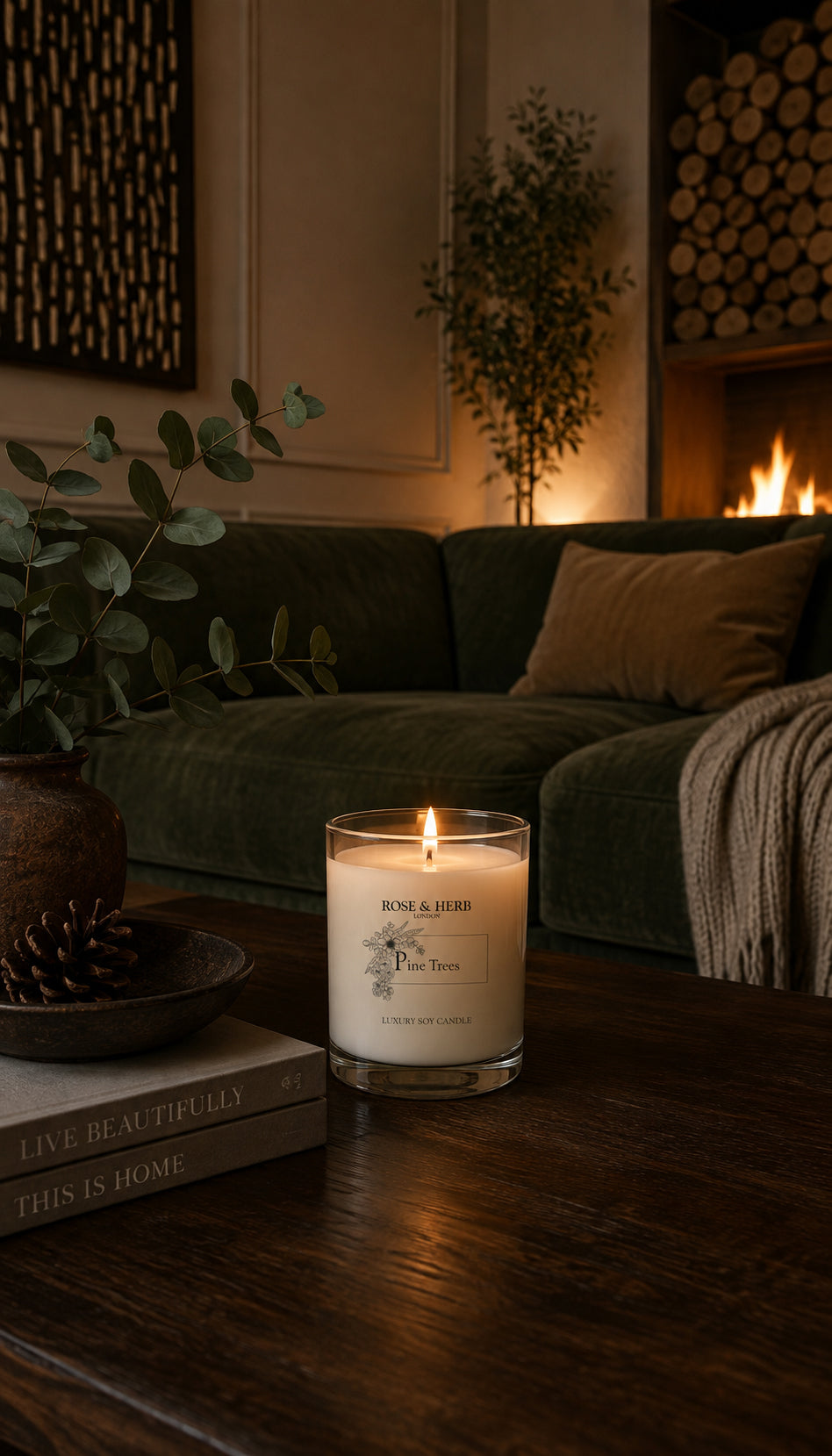 A lit soy candle in a clear glass jar sits on a dark wooden coffee table in a cozy living room setting with a green sofa, fireplace, and plants.