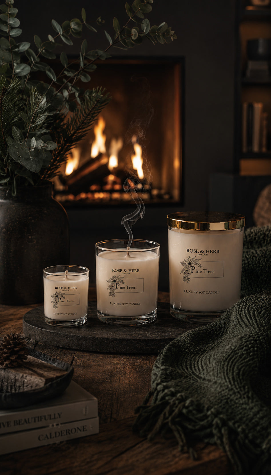 Three Rose & Herb scented candles in clear glass jars with white wax and gold lids, arranged on a wooden tray in front of a lit fireplace with a cozy green blanket nearby.