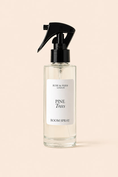 A clear glass room spray bottle with a black trigger spray top and a white label reading 'PINE Trees ROOM SPRAY' and 'ROSE & HERB LONDON', centered on a warm beige background.