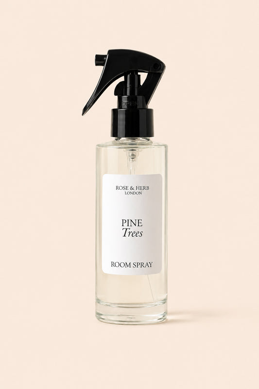 A clear glass room spray bottle with a black trigger spray top and a white label reading 'PINE Trees ROOM SPRAY' and 'ROSE & HERB LONDON', centered on a warm beige background.