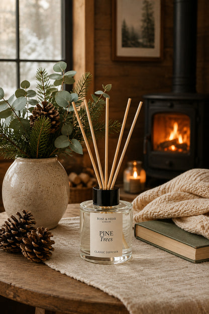 A clear glass reed diffuser bottle labeled Pine Trees with natural reed sticks, styled on a rustic wooden table beside a ceramic vase with greenery, pinecones, a candle, a knitted blanket, and a book in a cozy room with a fireplace.