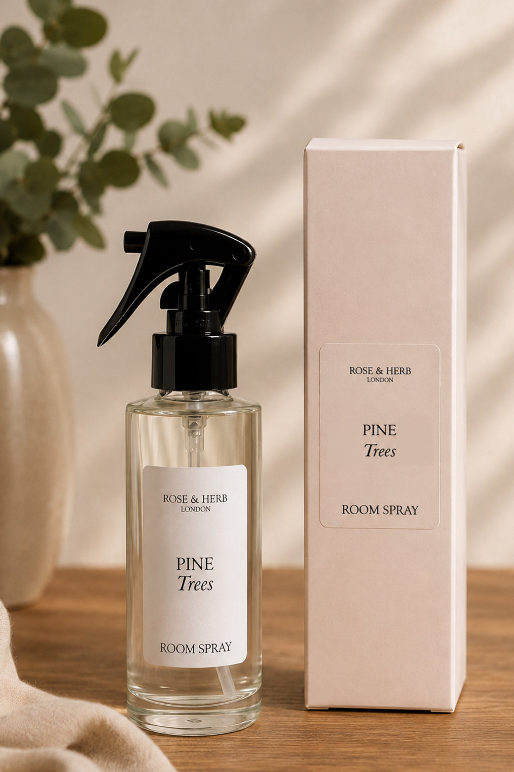 A clear glass room spray bottle with a black spray top and white label stands beside its white box on a wooden surface. The label reads 'ROSE & HERB LONDON' and 'PINE Trees ROOM SPRAY'.