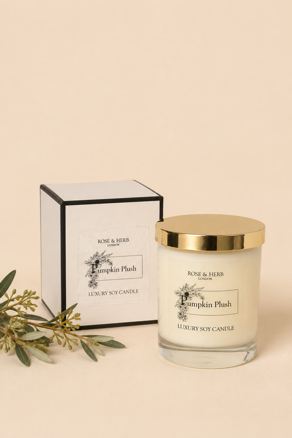 A luxury soy candle in a clear glass jar with a shiny gold lid, shown next to its white and black packaging box. The candle is labeled 'Pumpkin Plush' by Rose & Herb London.