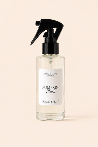 A clear glass room spray bottle with a black trigger spray top and a white label reading 'Rose & Herb London Pumpkin Plush Room Spray', centered on a warm beige background.