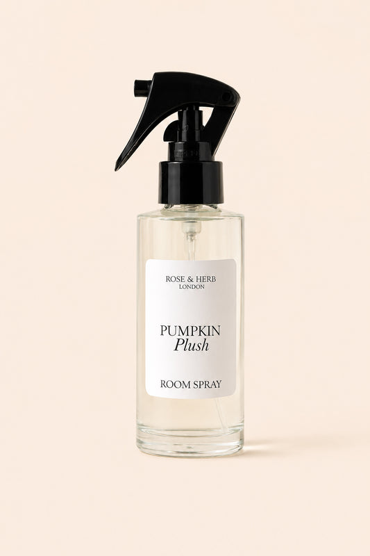 A clear glass room spray bottle with a black trigger spray top and a white label reading 'Rose & Herb London Pumpkin Plush Room Spray', centered on a warm beige background.